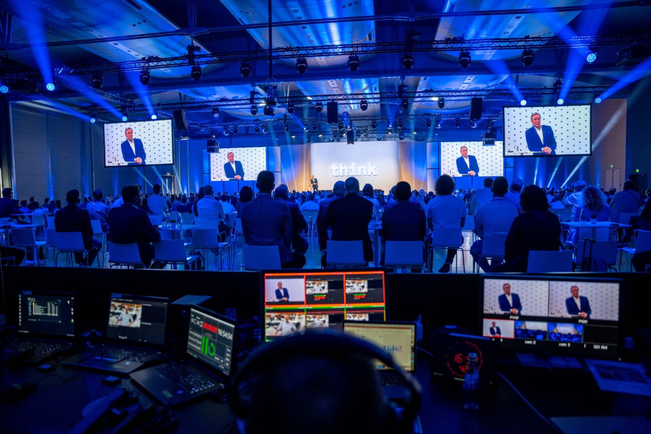 Einblick hinter die Kulissen der IBM Think on Tour. Video und Streamingequipment im Vordergrund. Im Hintergrund sind bühne, Publikum und Speaker zu sehen.